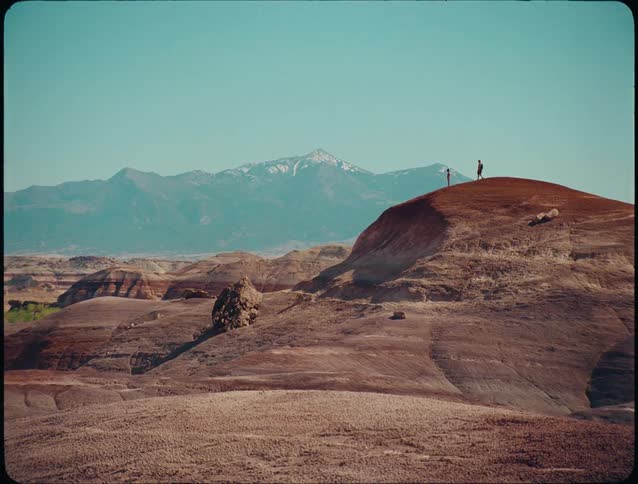 A wide view of a vast arid landscape shows two people standing atop a reddishbrown desert hill with distant snowcapped mountains visible under a clear blue sky
