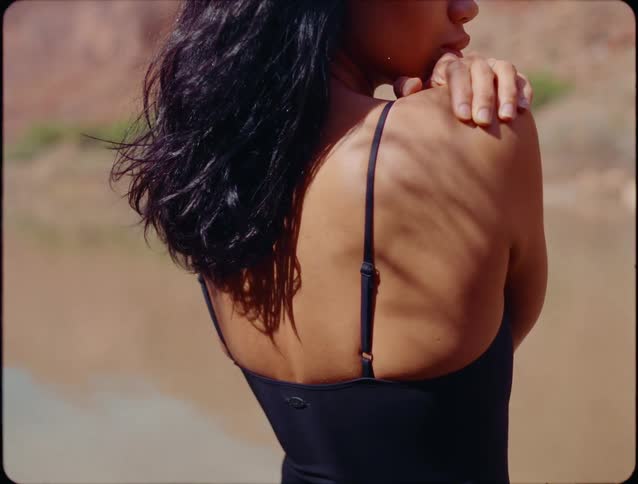 A close view of a woman with long dark hair wearing a black spaghetti strap top standing outside with her hand on her shoulder against a blurred sandy backdrop
