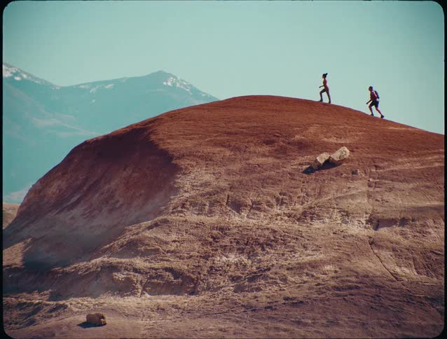 Two people climbing a reddish hill with mountains in the background under a clear sky
