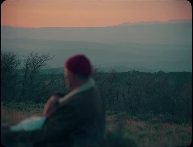 A man wearing a red beanie and a brown jacket sits outside holding a woman who is resting her head on his shoulder overlooking a vast dark landscape at sunset