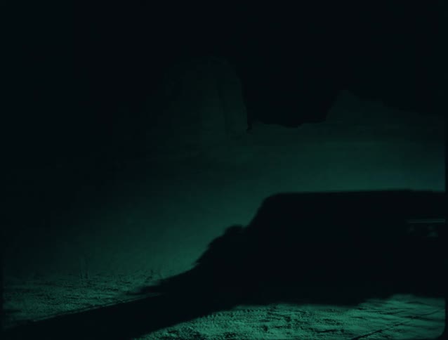 A car drives along a dark lonely road at night its headlights cutting through the quiet shadows as the surrounding landscape fades into silence