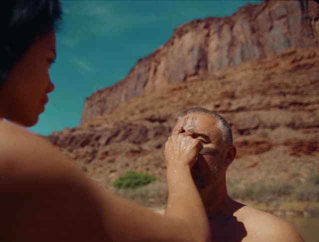 A medium outdoor view shows a woman with dark hair painting a white stripe of clay or pigment onto a mans forehead as they stand in a river with red canyon walls in the background