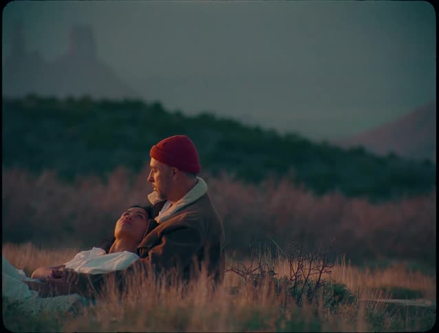 A young couple lies on a sunlit grassland at midday with the woman resting on the mans arm as the grasses around them fade to brown in the warmth of the fall season