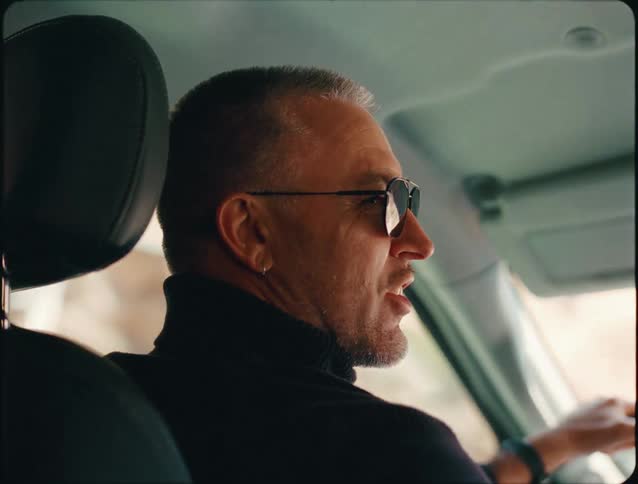 A close view over the passenger seat shows a smiling middleaged man with a beard and dark sunglasses driving a car while wearing a black turtleneck shirt