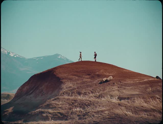 Two people walking on top of an arid hill with mountains in the background