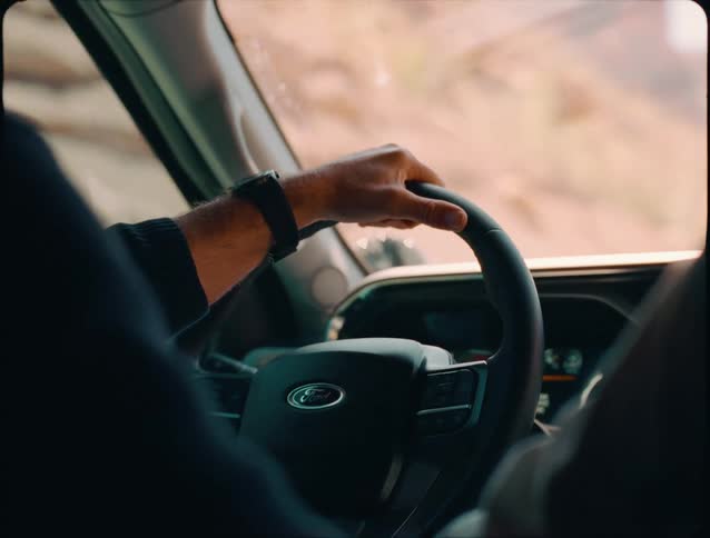 A close internal view shows a mans hands one wearing a dark watch gripping the ford steering wheel while driving offroad in a desert or dusty landscape