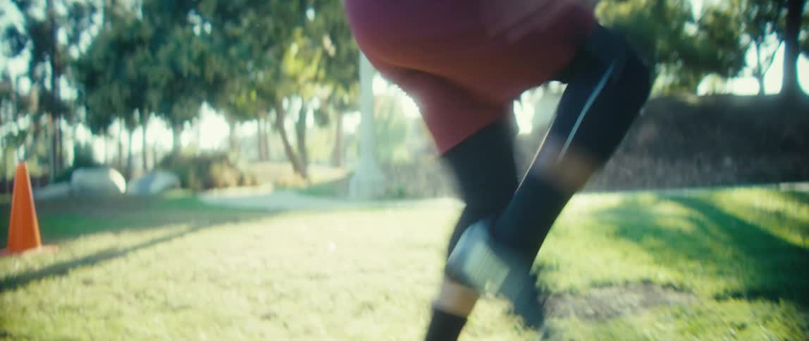 A young black male jogs in place in a sunlit public park surrounded by trees and open space as the midday light casts crisp shadows across the ground