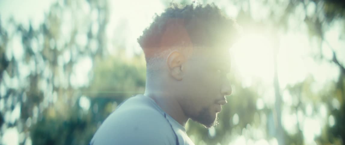 The video is a mediumcloseup profile shot of a man possibly an athlete with his head lowered in concentration or exhaustionthe scene is shot outdoors with a strong lens flare from the bright sunlight breaking through the blurry green tr