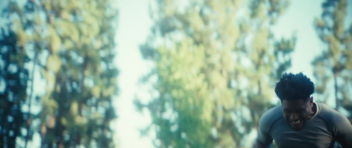 A young man runs through a sunlit public park with determination his stride focused as trees line the path and midday light glints off his activewear