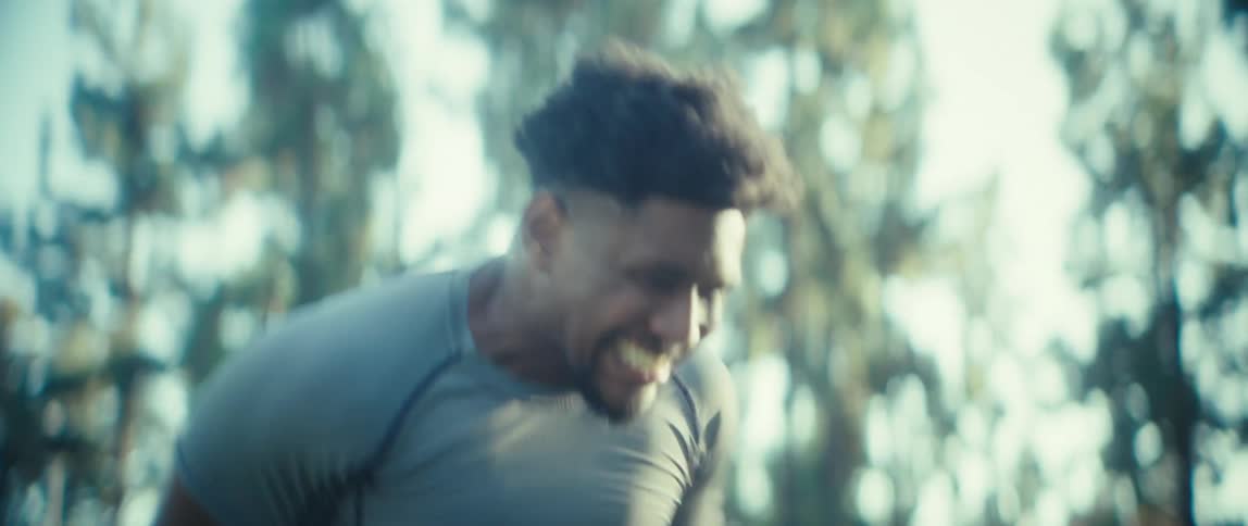 A young man jogs in place on a sunny day in a public park surrounded by trees and open space warm light highlights his focused movement steady rhythm and determination