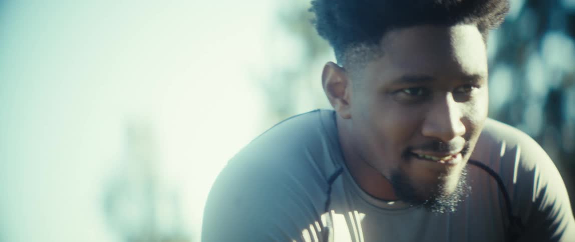 A young black man in activewear squats in a public park on a sunny day catching his breath and looking tired after a workout