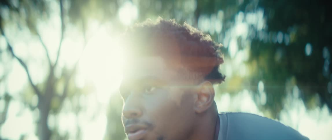 A black male who appears tired has his hands on the back of his head then leans forward near trees