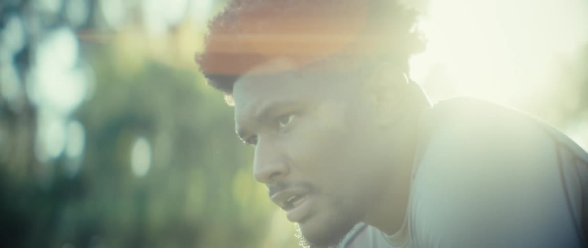 A closeup shot focuses on the face and upper torso of a man with dark curly hair and a beard as he breathes heavily while bent over in a posture of physical exhaustion with bright sunlight creating a strong lens flare in the outdoor setting