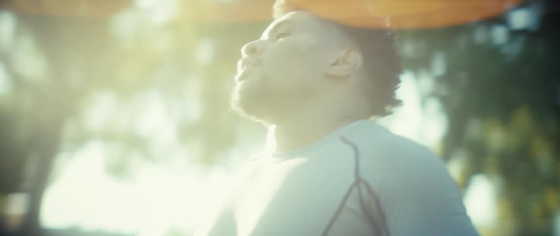 A close view of a man with an afro hairstyle and a beard wearing a gray athletic shirt wiping his neck and face with a white towel outdoors after a workout in bright sunlight