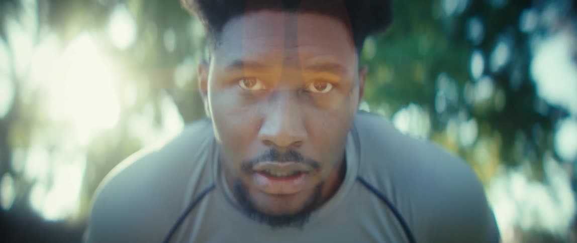A close lowangle view captures a man with an afro hairstyle and a beard wearing a gray athletic shirt pausing to breathe heavily after a workout backlit by bright sun and lens flares