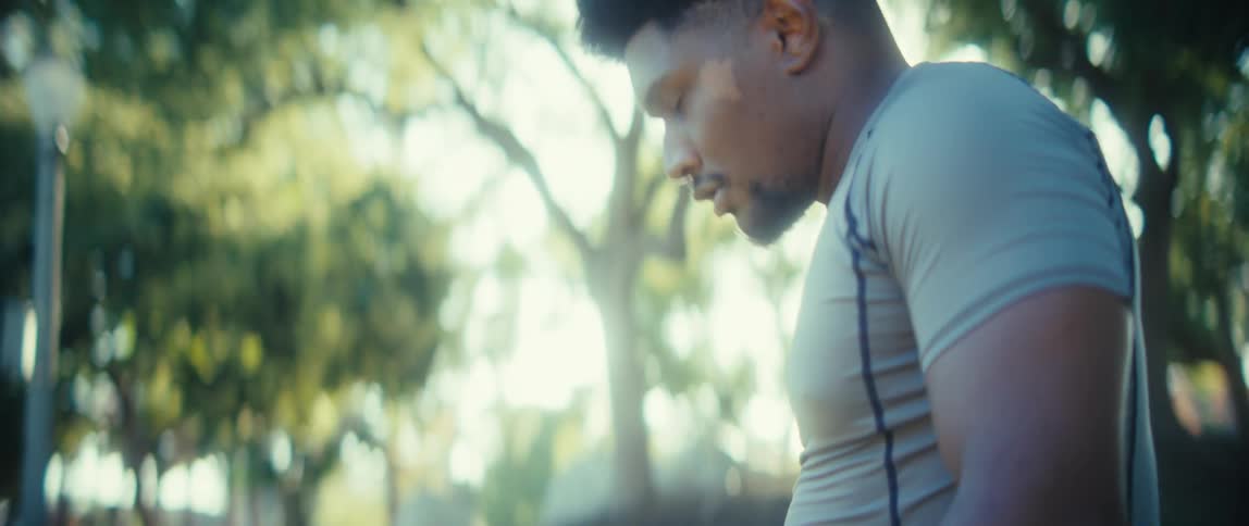 A young man jogs in place on a sunny day in a public park surrounded by trees and open space as warm light highlights his focused movement and steady rhythm