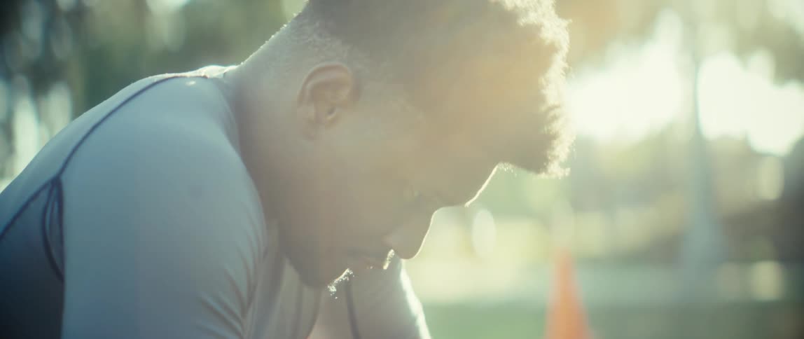 A young black man sits under the sun in a public park looking tired after a workout