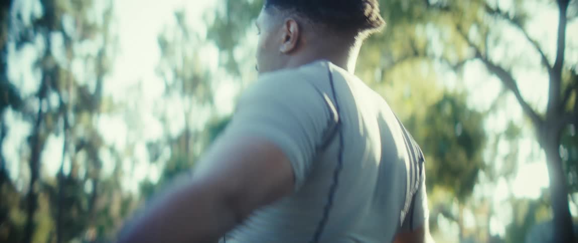 The video clip shows an athletic man exercising outdoors in a parklike setting with rows of trees in the background he is wearing a grey shirt and red shorts and is performing a side bend stretch with his arms clasped above his head