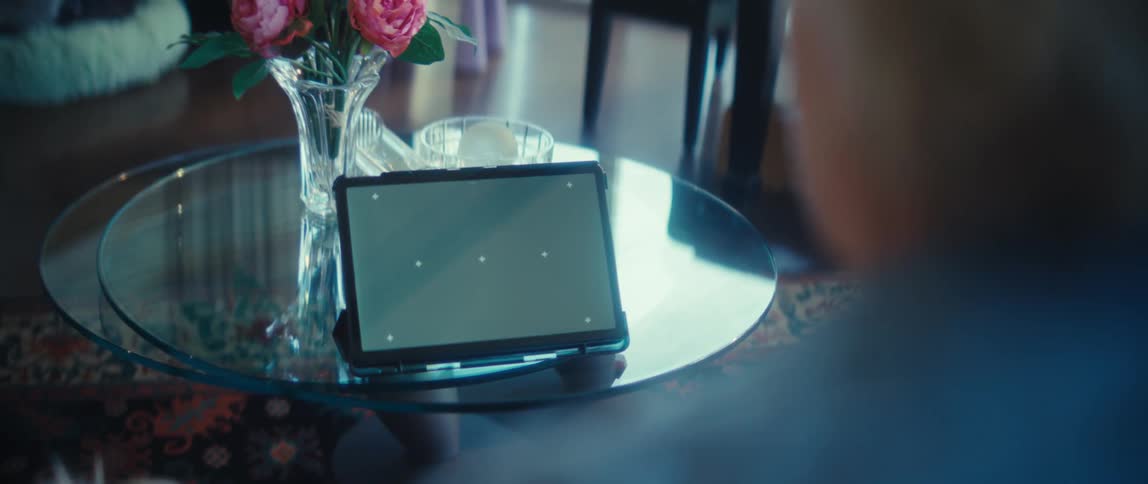 A person watches a tablet with a green screen propped up on a glass table next to a vase of flowers
