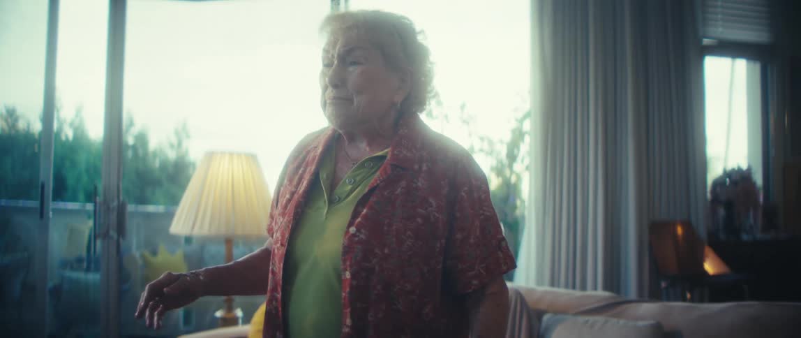 A wide bright interior view shows an older woman in a green shirt and patterned red blouse dancing by a couch in her living room singing heartily by a large sunlit window