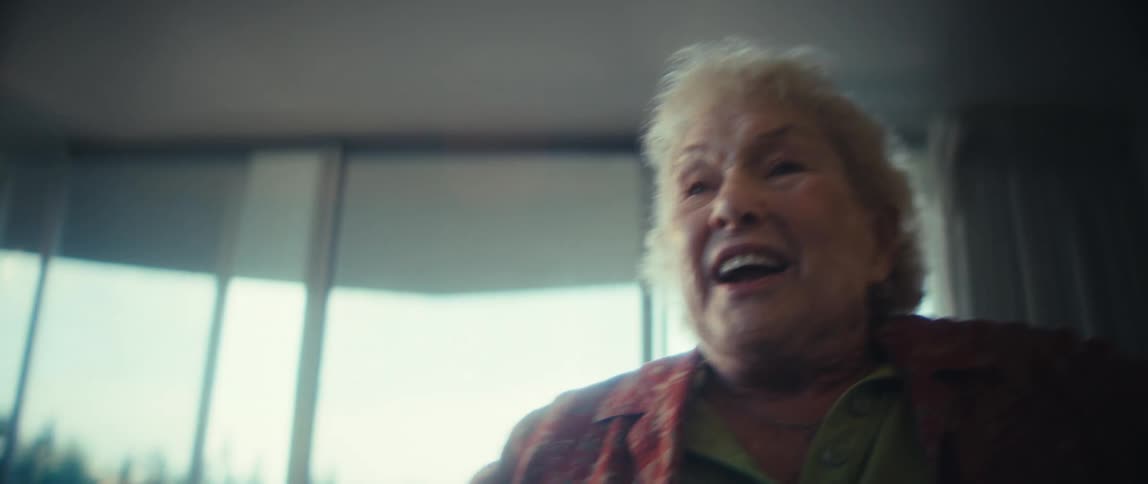 A smiling and talking older woman with curly blond hair stands indoors by a bright window in a floral blouse