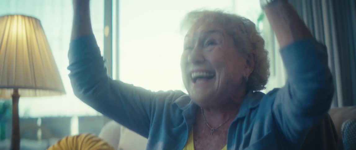 A close view shows a very happy older woman in a blue shirt and yellow top cheering and raising her arms in excitement while sitting in a brightly lit living room