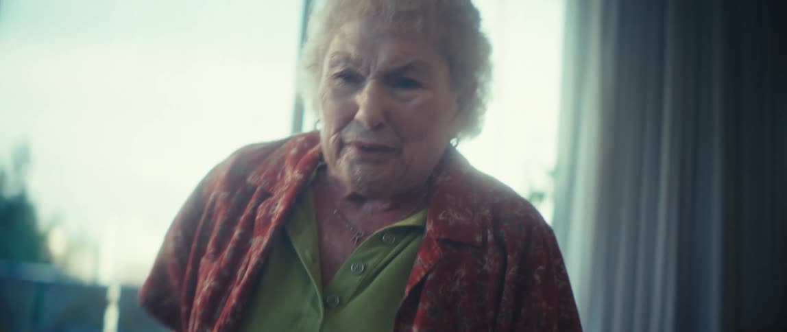 A close view of a cheerful older woman with blonde hair wearing a green shirt and red patterned blouse looking down and then smiling while gesturing excitedly in a bright room