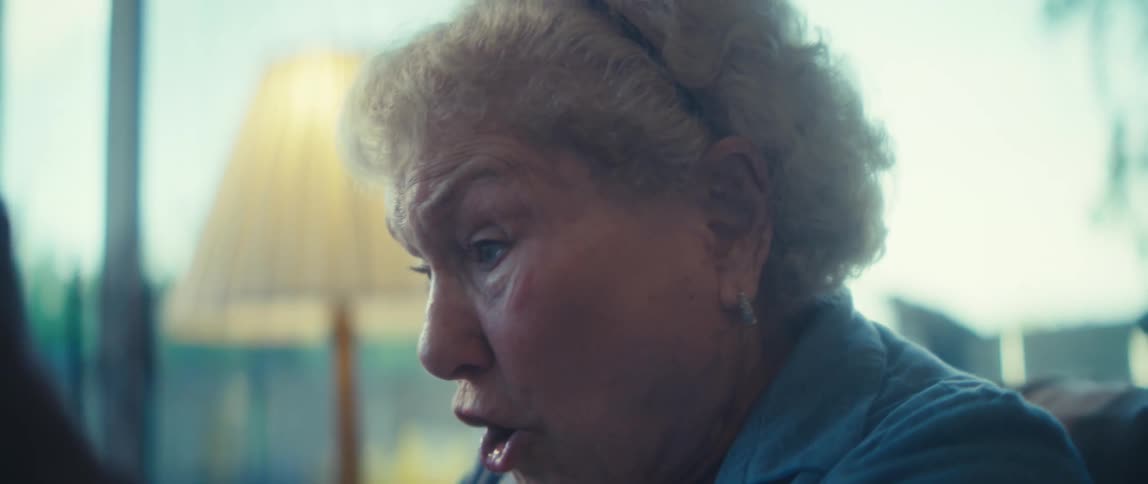 The video clip is a mediumclose shot of an elderly woman with short white hair expressing surprise or excitement her mouth is open in an o shape and she is wearing a light blue collared shirt and a delicate necklace