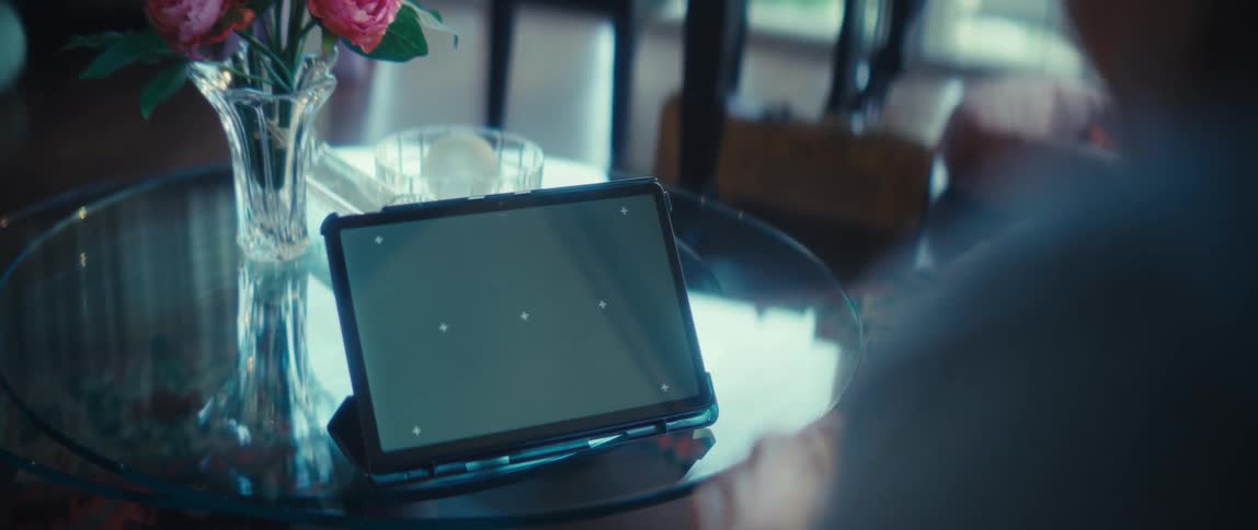An elderly woman interacting with a screen of a black tablet which rests on a glass table next to a vase of pink roses in a welllit indoor space