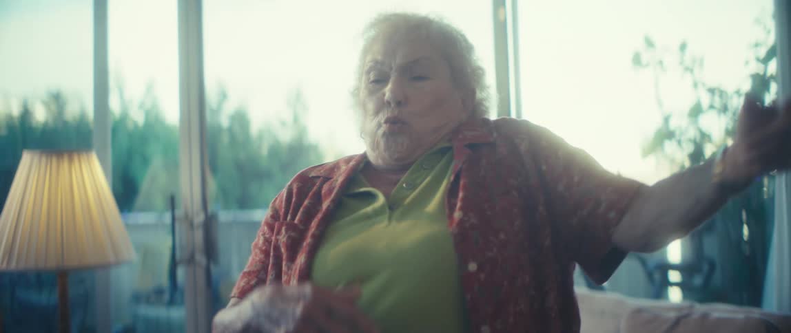 A wide brightly backlit view shows an older woman in a green shirt and patterned red blouse dancing enthusiastically in her living room by a large window