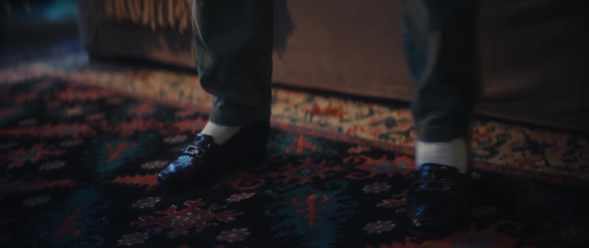 A close view shows a persons feet wearing black shiny loafers with white socks and dark pants tapping on an ornate patterned rug in a dim interior