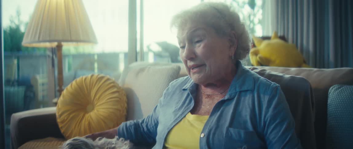 Elderly woman sitting on the couch with two dogs resting beside her while talking to someone on a tablet