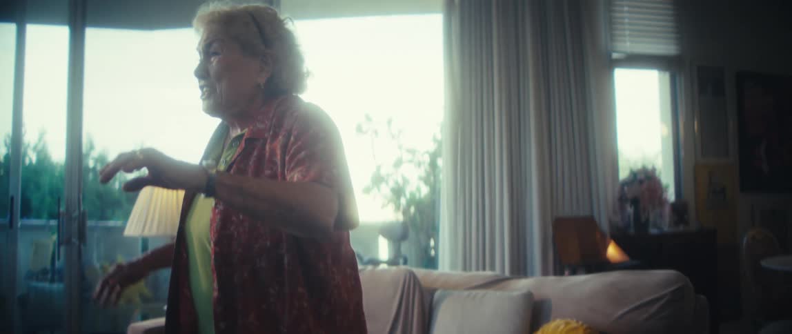 A full view shows a happy older woman in a red patterned shirt and green top dancing in her brightly lit living room with a small white dog resting on the couch behind her