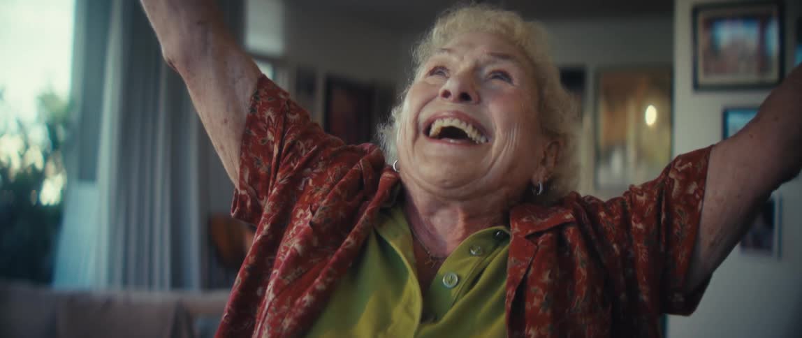 An elderly woman with white curly hair in a red patterned shirt raising both arms above her head and smiling widely with her head tilted upward in an expression of pure joy in a brightly lit indoor space