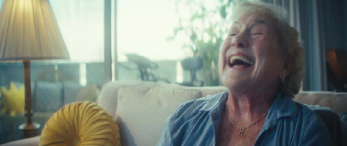 A full view of a joyful older woman in a yellow shirt and blue button down shirt sitting on a couch with two fluffy white dogs cheering with her hands up in front of a bright window