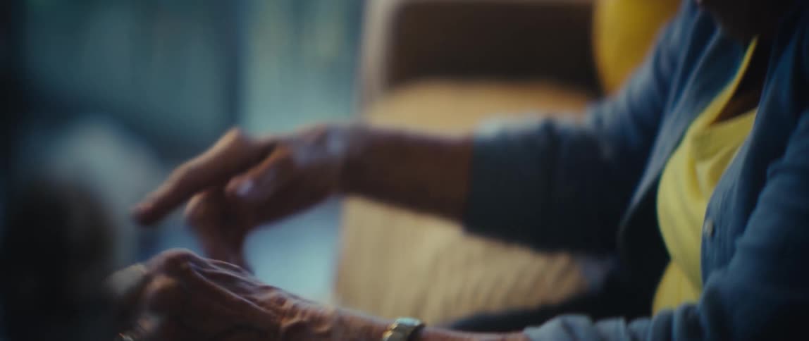 An elderly persons hands gently petting and stroking the curly white head and neck of a small dog that is looking up at the person while they sit together in an indoor setting