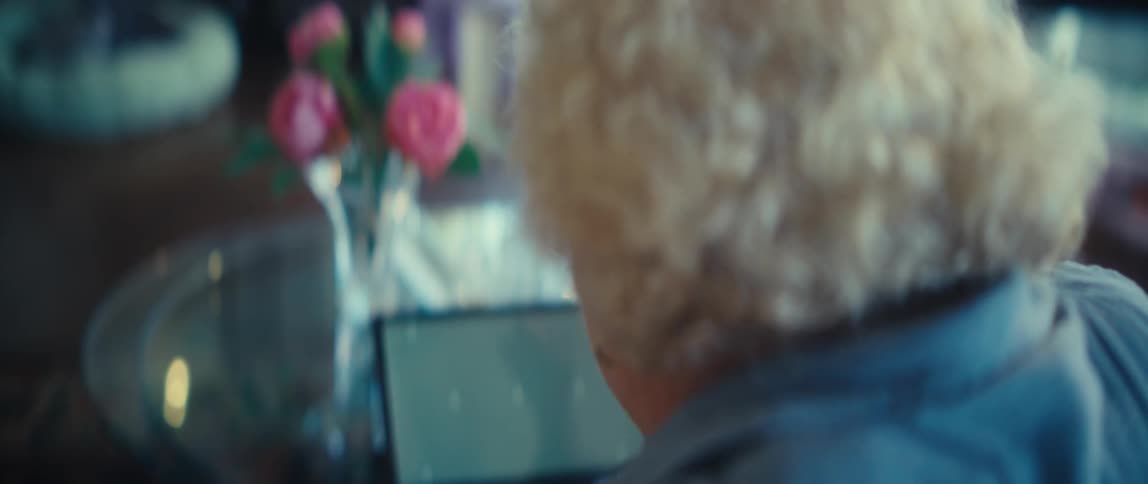 An elderly woman with white curly hair sitting at a glass table and operating a small black tablet in a living room setting