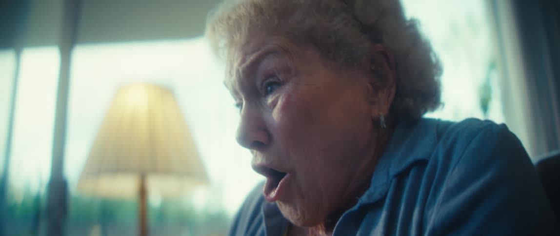 A close view of a joyful older woman in a yellow shirt and blue buttondown clapping and cheering happily in a brightly lit living room with a fluffy white dog resting on the couch