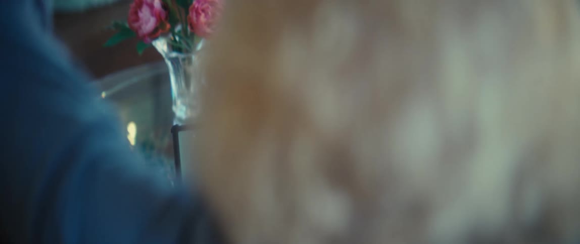 A blurry overtheshoulder view shows an older woman with blonde hair using a tablet with a green screen on a glass table next to a vase of pink flowers