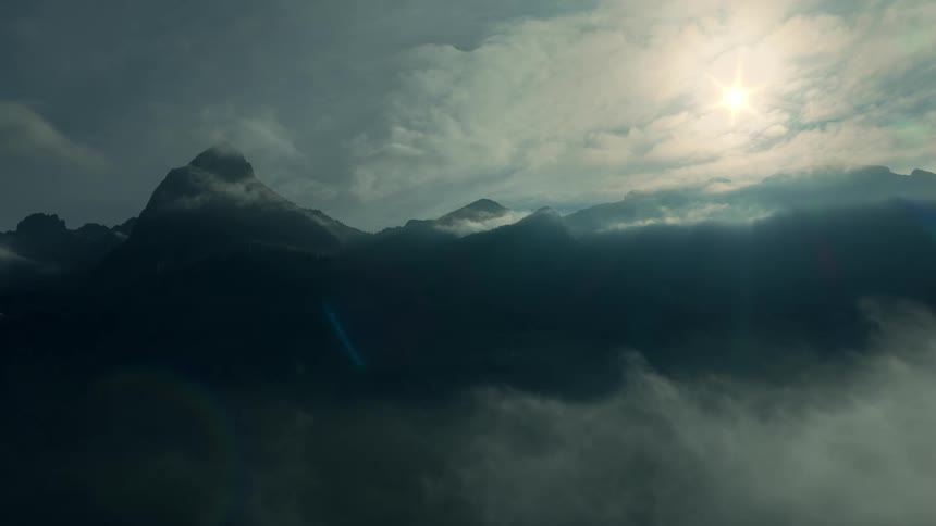 Dark silhouette of mountain peaks partially obscured by low moving clouds or mist below with bright sunlight breaking through the cloudy sky above and creating a lens flare