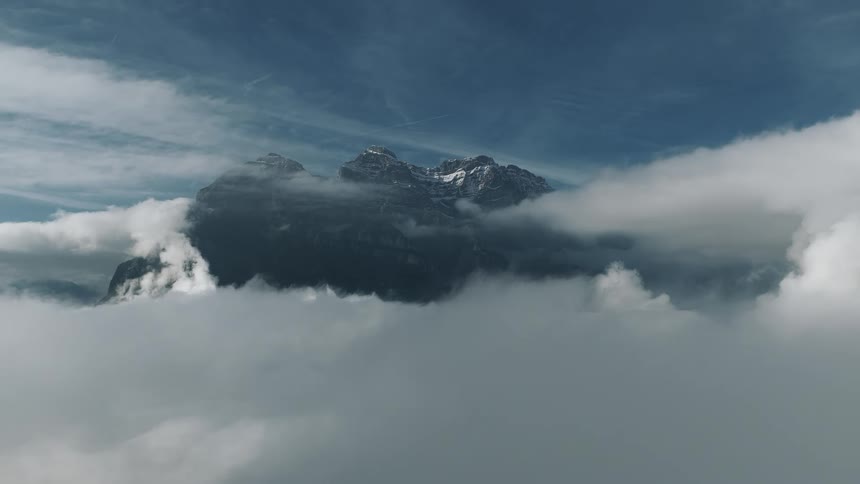 A dramatic aerial view shows dark jagged alpine peaks with patches of snow rising high above a layer of thick fluffy white clouds under a deep blue sky