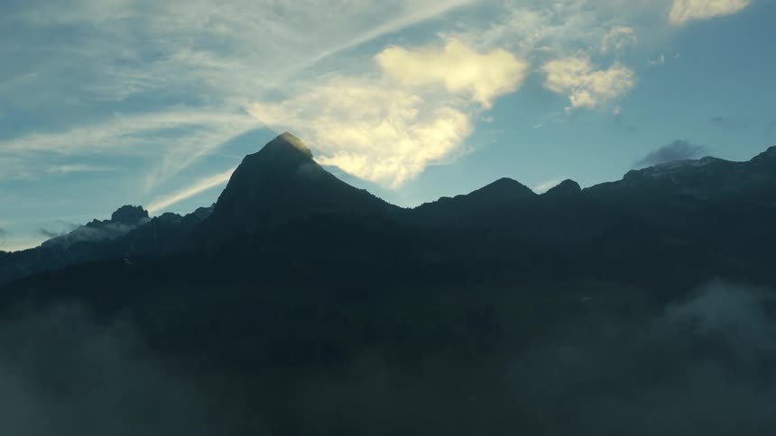 A lowangle wide shot shows dark jagged alpine peaks with the central peak dramatically backlit by glowing sunlight behind wispy clouds in a blue sky