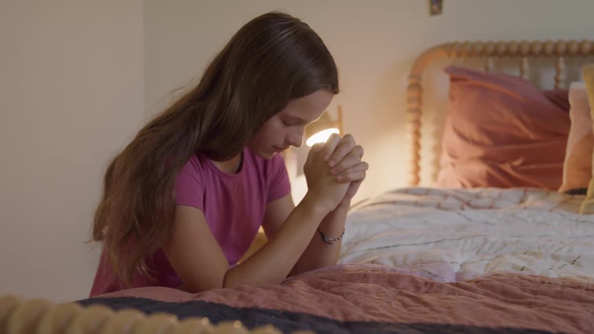 A young girl with long dark hair kneeling or sitting on a bed with her head bowed and her hands clasped together under her chin in a posture of prayer in a softly lit bedroom