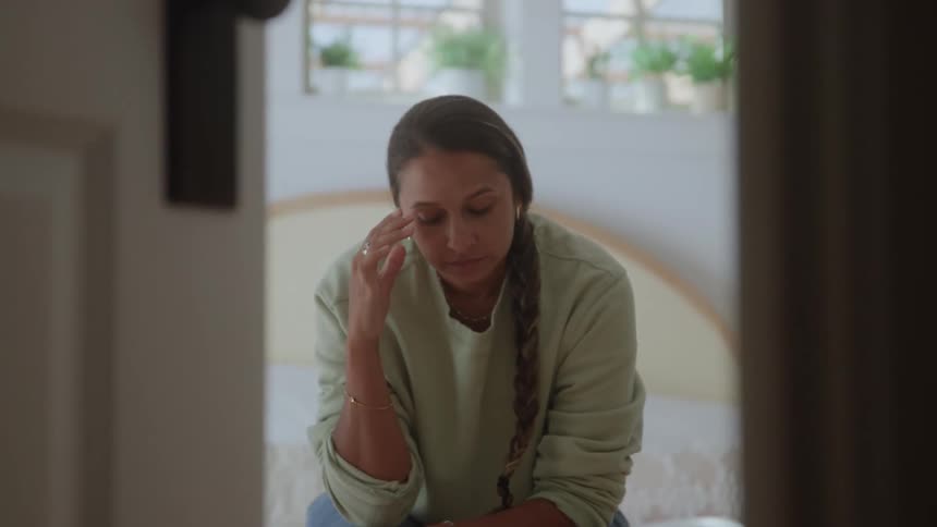 A woman with a long braid sitting and leaning forward with her eyes closed touching her forehead with one hand in a gesture of distress or pain in a brightly lit room