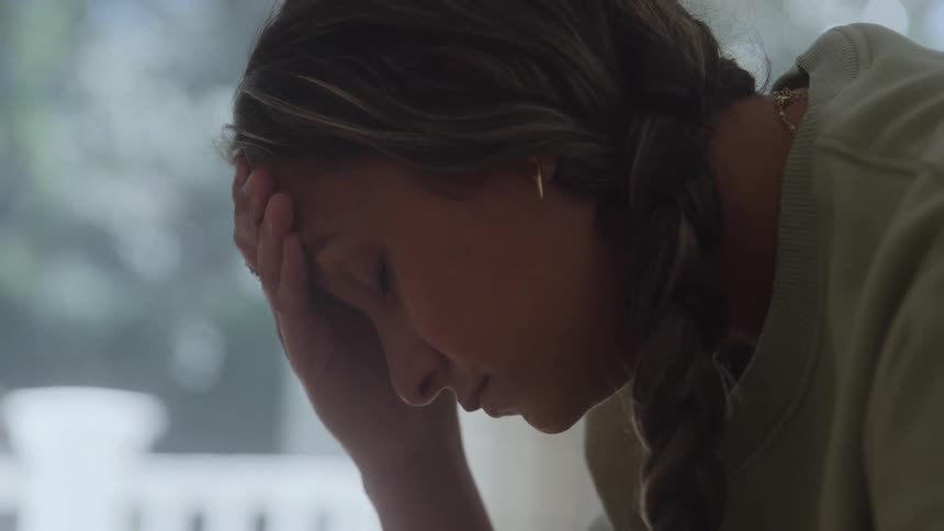 A young woman illuminated by soft window light rests her head on her hand with a look of contemplation or stress