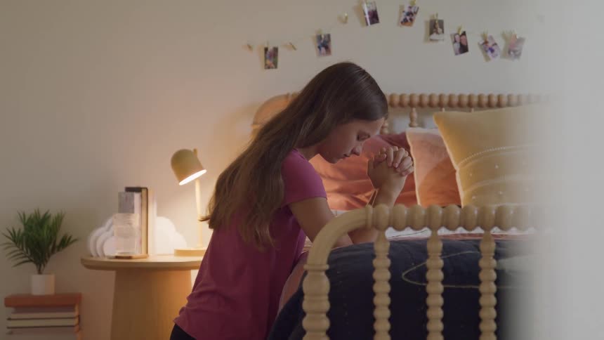 A female teenager praying while kneeling beside her bed in her bedroom during daytime