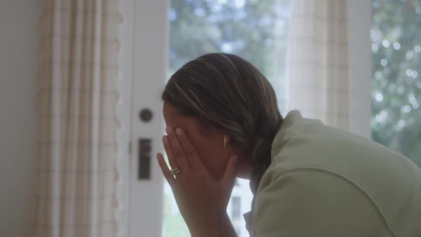 A woman indoors covers her face with both hands near a bright window suggesting stress or overwhelm