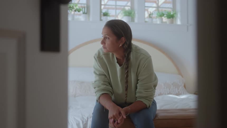 A woman in a green sweatshirt sits on a bench at the edge of a bed with hands clasped gazing toward the bright window thoughtfully