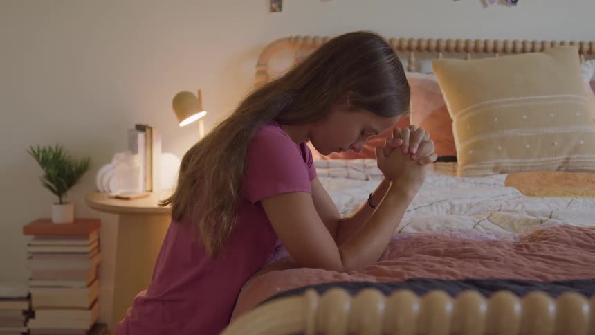 A young female in a pink shirt kneels by a cozy bed with clasped hands and a bowed head in quiet prayer