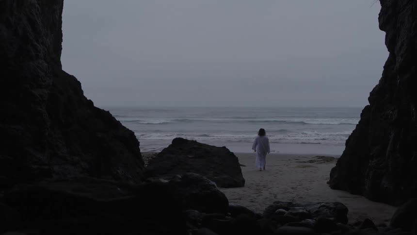 The video clip shows a distant view of a person wearing a white robe walking toward the ocean on a dark beach the scene is framed by dark rock formations on both the left and right creating a natural archway effect